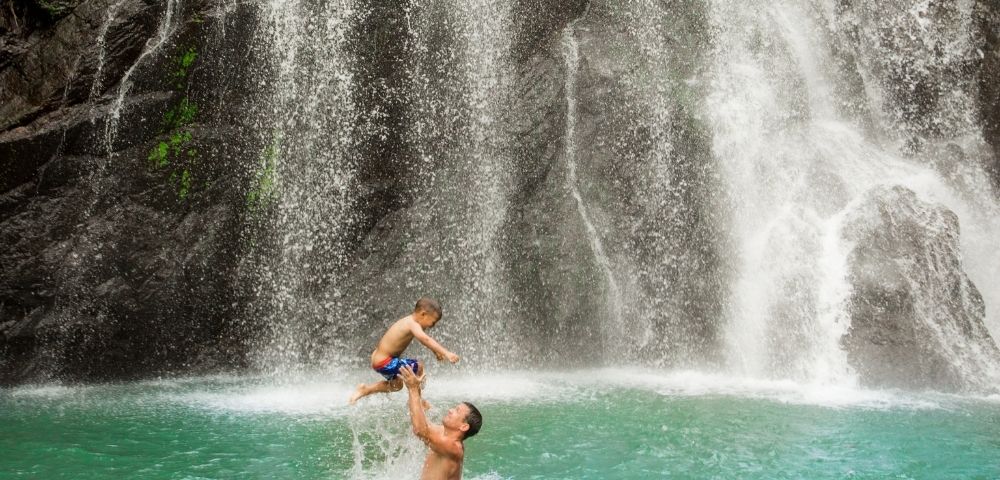 father throwing his son up in the air while playing in the water in front of a waterfall