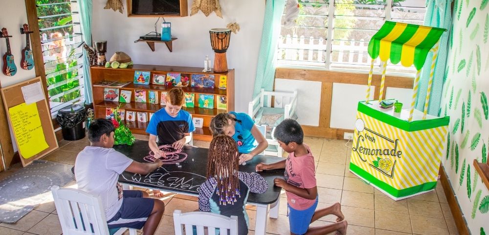 five children drawing on a blackboard table with a play lemonade stand in the background