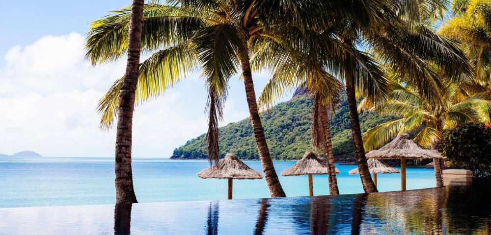 Pool with palm trees overlooking beach with straw top wooden umbrellas