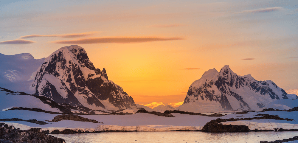 Sunset in a polar landscape with snow-covered mountains and penguins near water.