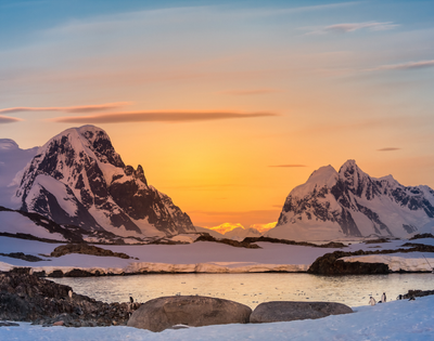 Sunset in a polar landscape with snow-covered mountains and penguins near water.