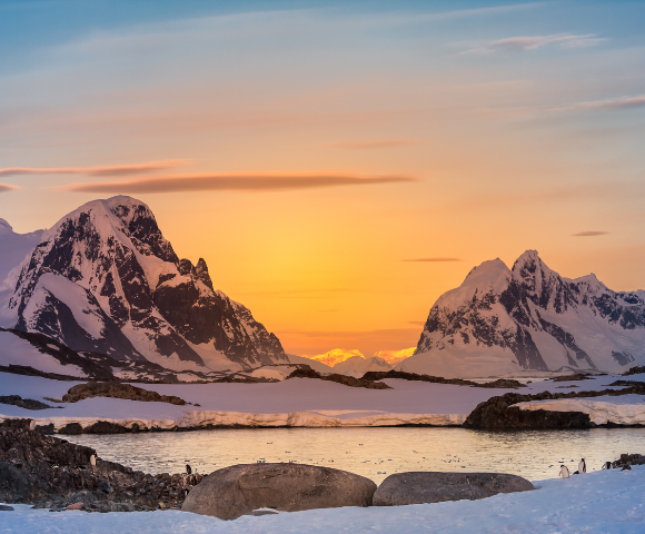 Sunset in a polar landscape with snow-covered mountains and penguins near water.