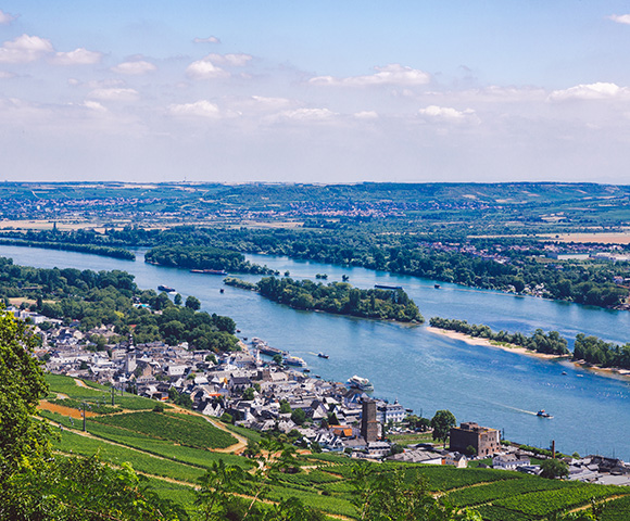 Aerial view of a river with boats, towns, vineyards, and green hills under a partly cloudy sky.