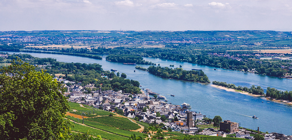 Aerial view of a river with boats, towns, vineyards, and green hills under a partly cloudy sky.