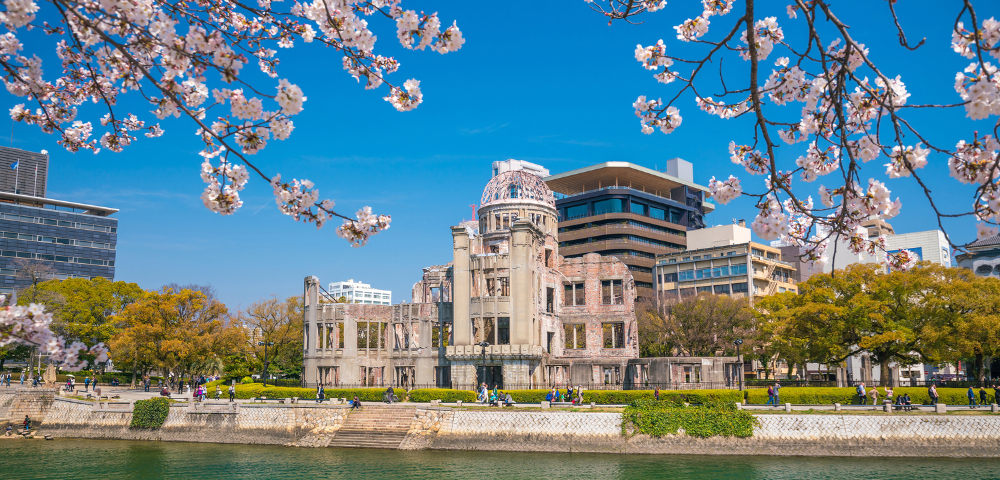 Cherry blossoms frame a riverfront view of a historical building with modern buildings behind it under a blue sky.