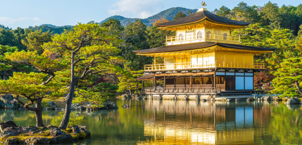 Golden pavilion reflecting on a pond surrounded by trees under a clear sky.