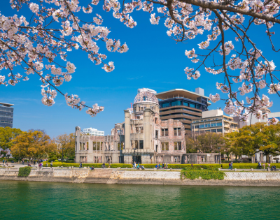 Blossoming cherry trees frame a view of a riverfront with classical and modern buildings under a clear blue sky.