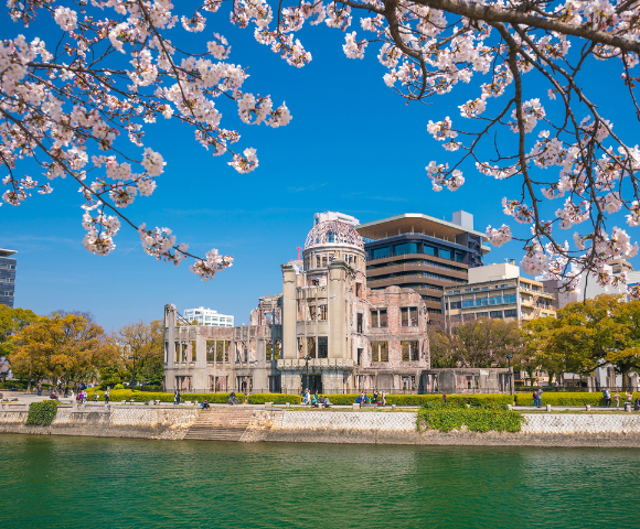 Cherry blossoms frame a view of the Hiroshima Peace Memorial by a river under a blue sky.
