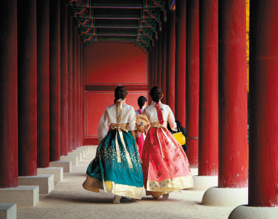 Three individuals in traditional Korean hanboks walking through a red-pillared corridor.