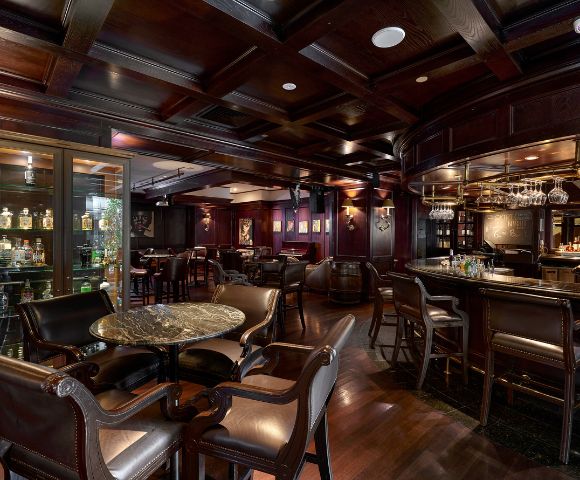 Interior of a moody bar with dark wood burgundy walls and marble tables with a range of spirits in the cabinet