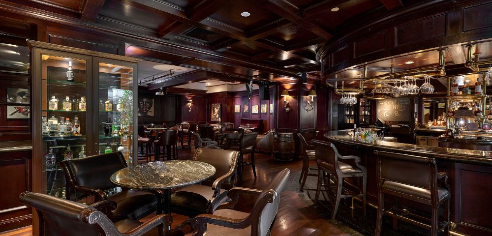 Interior of a moody bar with dark wood burgundy walls and marble tables with a range of spirits in the cabinet