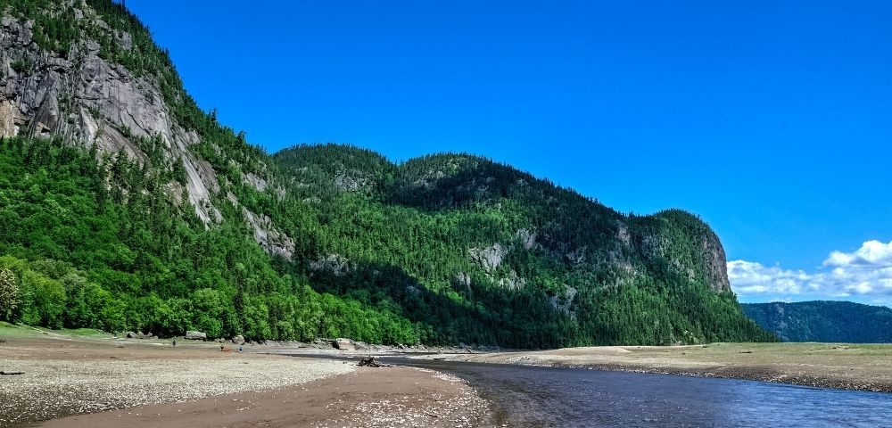 River winding through a sandy shore with forest-covered cliffs under a blue sky.