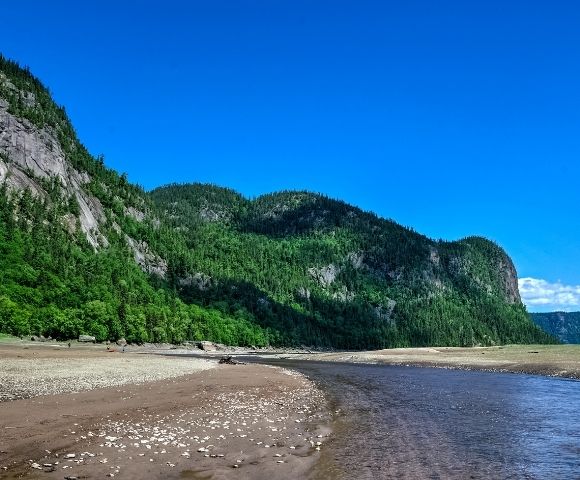 River winding through a sandy shore with forest-covered cliffs under a blue sky.