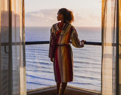 A person in a colorful robe standing on a balcony overlooking the sea at sunset.