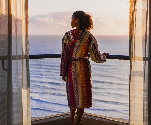 A person in a colorful robe standing on a balcony overlooking the sea at sunset.
