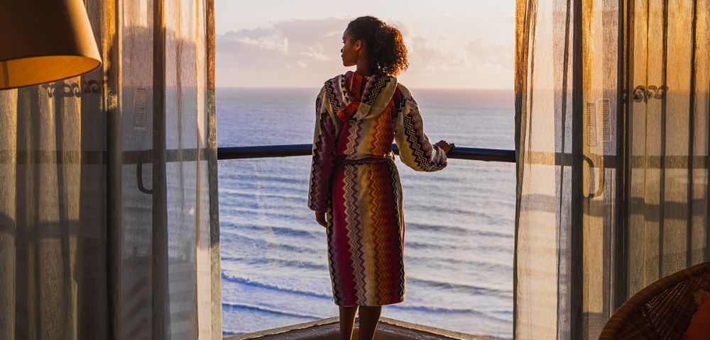 A person in a colorful robe standing on a balcony overlooking the sea at sunset.