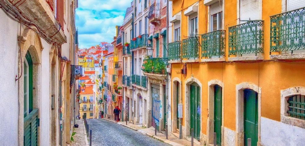 Colorful buildings line a narrow cobblestone street in an old European town, under a blue sky with clouds.