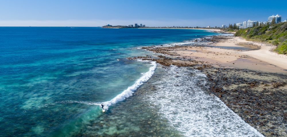 Coastal view with clear blue water, waves hitting rocky shore, and beach extending towards a distant city.