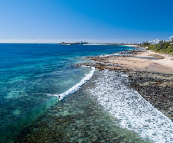 Coastal view with clear blue water, waves hitting rocky shore, and beach extending towards a distant city.
