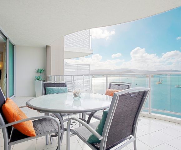 Balcony with a table and chairs overlooking a calm sea with boats and mountains in the distance.