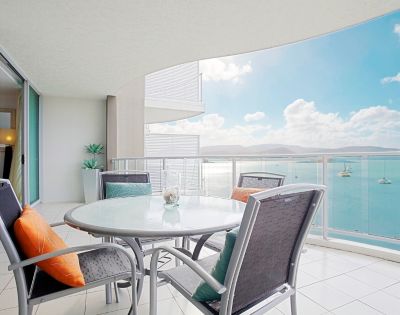 Balcony with a table and chairs overlooking a calm sea with boats and mountains in the distance.