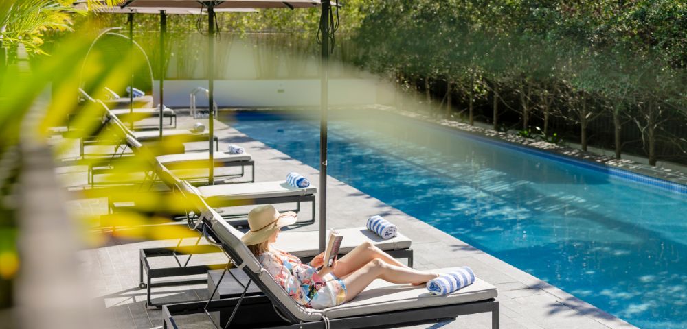 Woman relaxing on a sun lounger by a pool, reading a book under an umbrella.