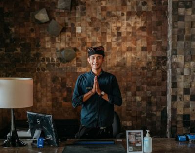 Person with hands pressed together in a gesture of respect, standing by a lamp and laptop against a textured wall.