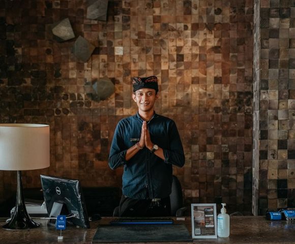 Person with hands pressed together in a gesture of respect, standing by a lamp and laptop against a textured wall.