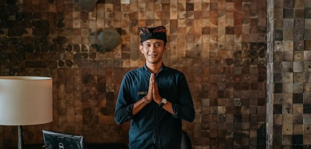 Person with hands pressed together in a gesture of respect, standing by a lamp and laptop against a textured wall.