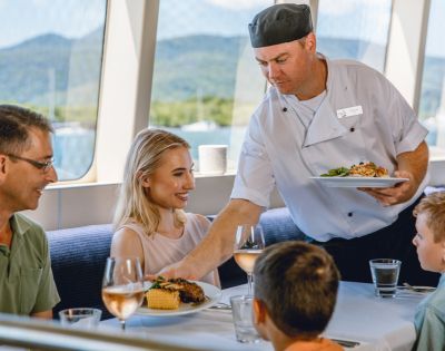Chef serving food to a family dining on a boat with scenic views through the windows.