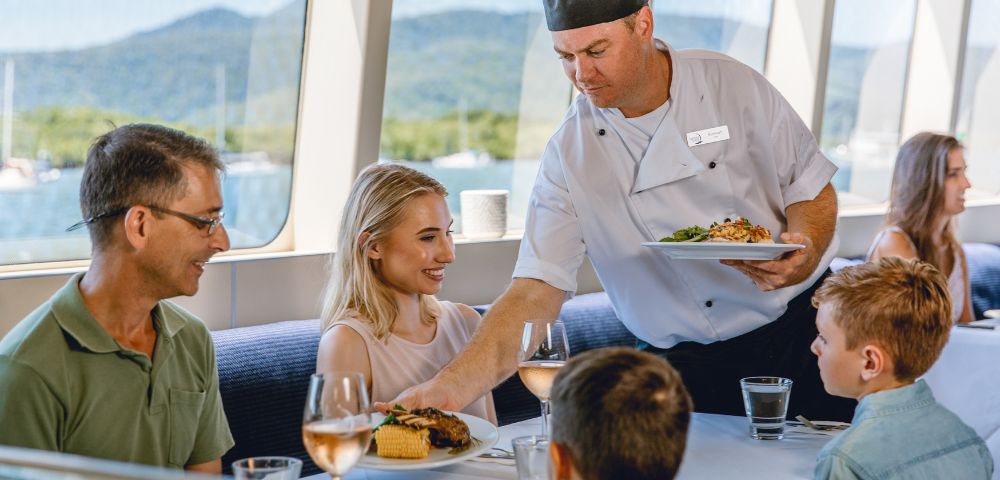 Chef serving food to a family dining on a boat with scenic views through the windows.