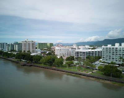 Aerial view of a coastal cityscape with modern buildings along a river, against a backdrop of mountains and clouds.
