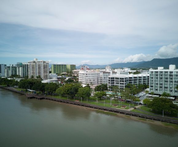 Aerial view of a coastal cityscape with modern buildings along a river, against a backdrop of mountains and clouds.