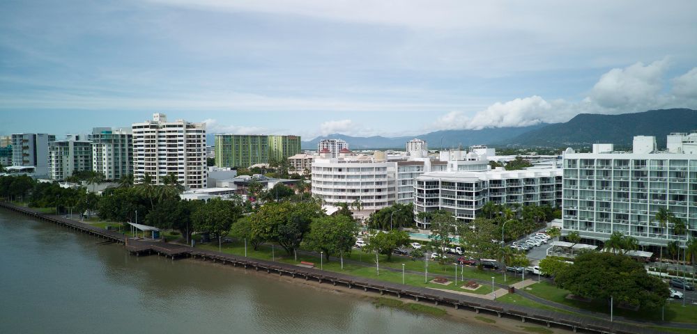 Aerial view of a coastal cityscape with modern buildings along a river, against a backdrop of mountains and clouds.