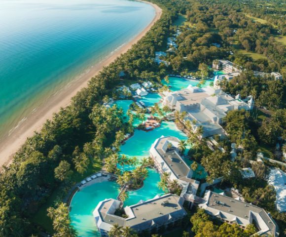 Aerial view of a tropical resort with pools by the beach, surrounded by lush greenery.