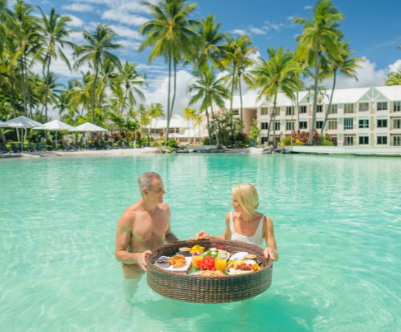 Couple having a floating breakfast in a tropical resort pool surrounded by palm trees.