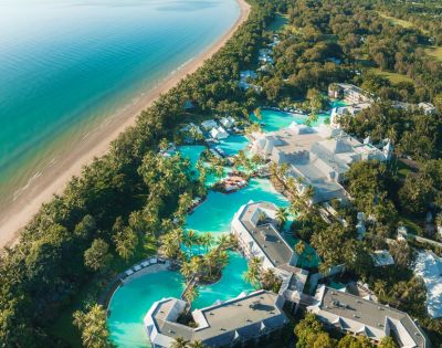 Aerial view of a tropical resort with pools by the beach, surrounded by lush greenery.