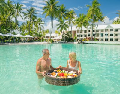 Couple having a floating breakfast in a tropical resort pool surrounded by palm trees.