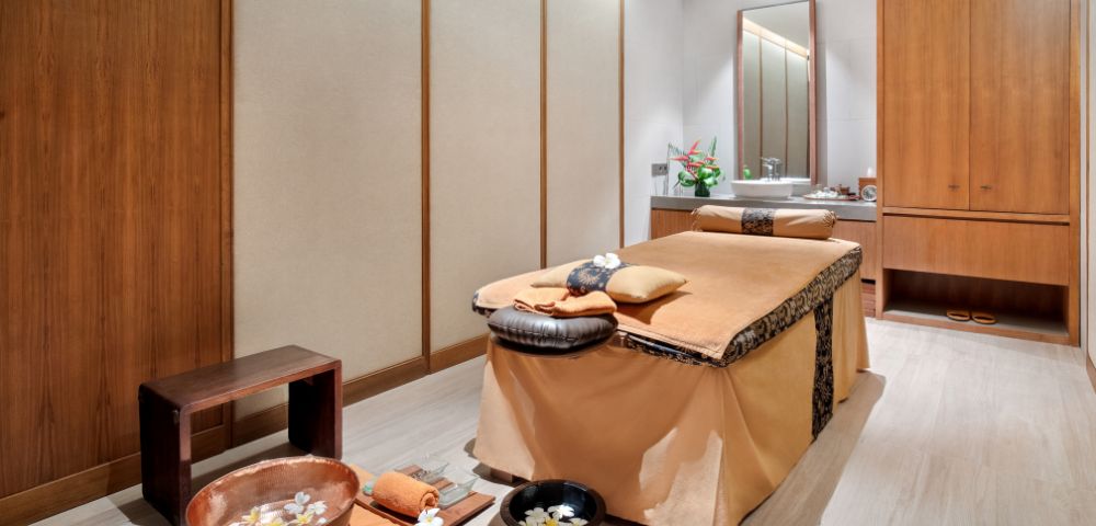 A serene spa room with a massage table, wooden accents, and bowls with flowers.