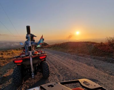 Rider on an ATV holding a flag, facing a sunset on a dirt road.