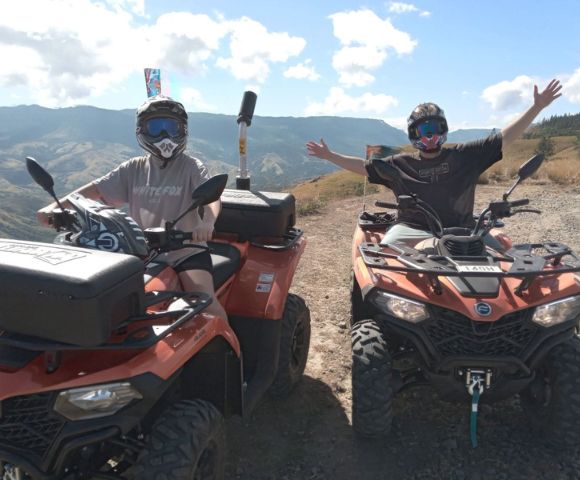 Two people on ATVs posing with outstretched arms on a mountain trail.