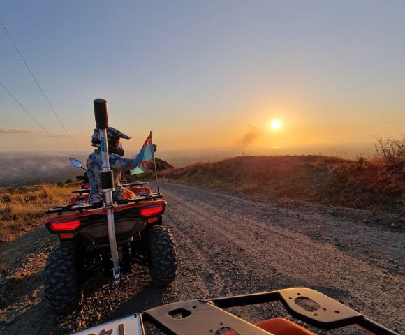 Rider on an ATV holding a flag, facing a sunset on a dirt road.