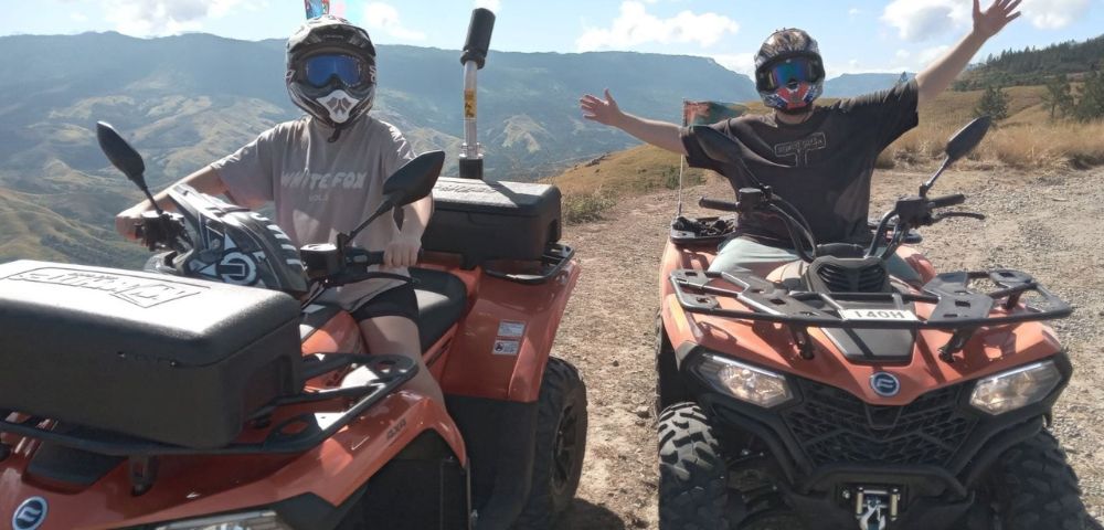 Two people on ATVs posing with outstretched arms on a mountain trail.