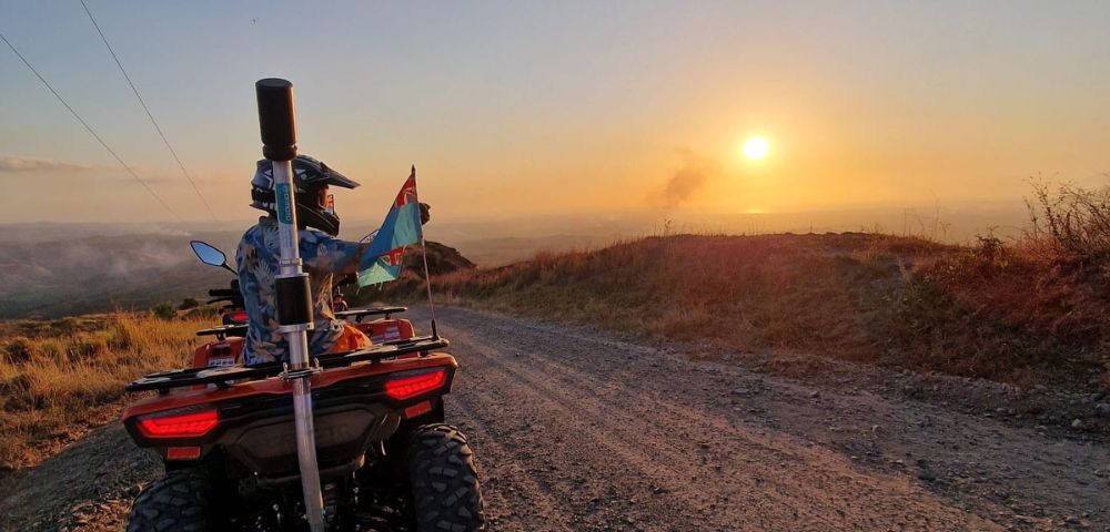 Rider on an ATV holding a flag, facing a sunset on a dirt road.