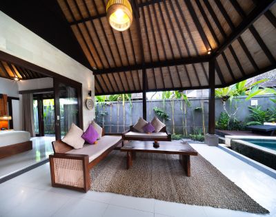 Open-concept living room with wooden furniture, high ceiling, and kitchenette, leading to a bedroom.
