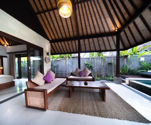 Open-concept living room with wooden furniture, high ceiling, and kitchenette, leading to a bedroom.