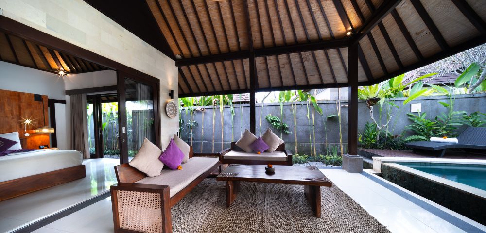 Open-concept living room with wooden furniture, high ceiling, and kitchenette, leading to a bedroom.