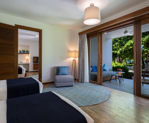 A modern hotel room with two beds, an armchair, and open doors leading to a balcony with greenery.