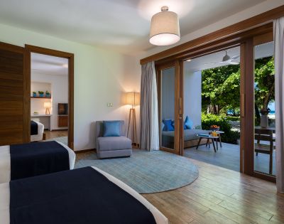 A modern hotel room with two beds, an armchair, and open doors leading to a balcony with greenery.