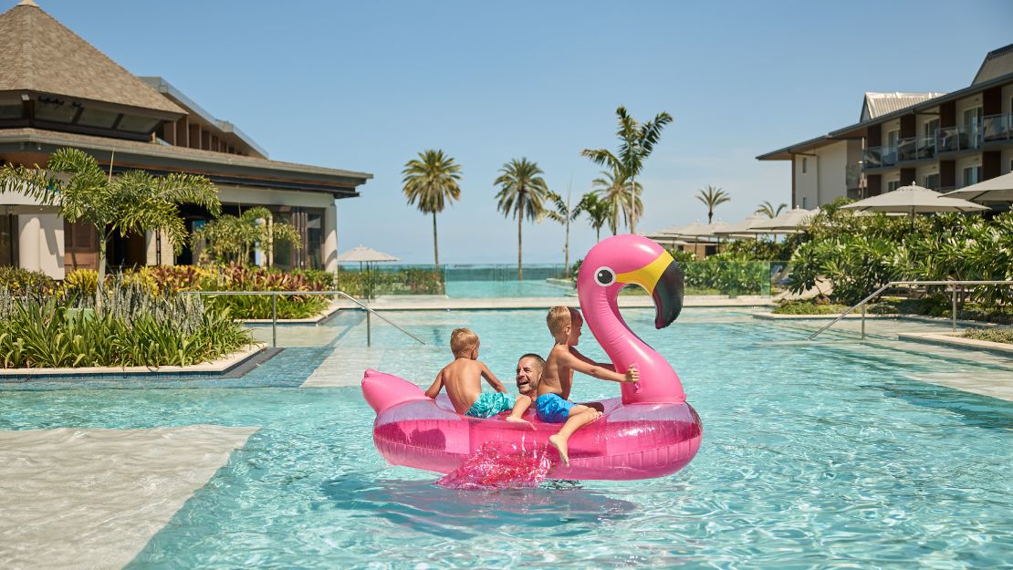 Crowne Plaza Fiji Nadi Bay Resort and Spa Two children on a pink flamingo float in a resort pool with palm trees and buildings in the background.
